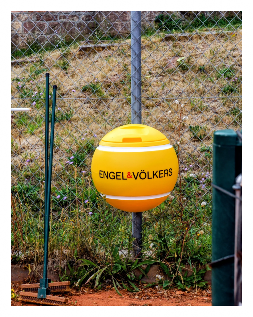 Gelber Tennisball-Mülleimer von Engel & Völkers am Zaun eines Tennisplatzes