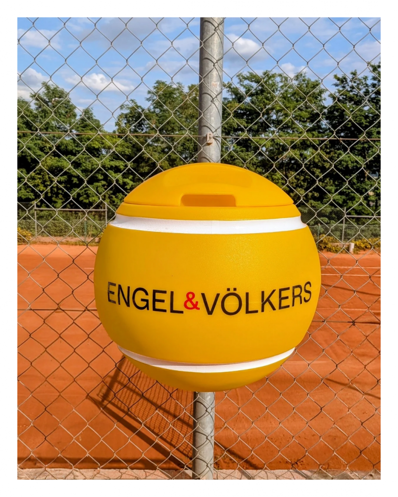Gelber tennisballförmiger Mülleimer von Engel & Völkers auf einem Sandplatz