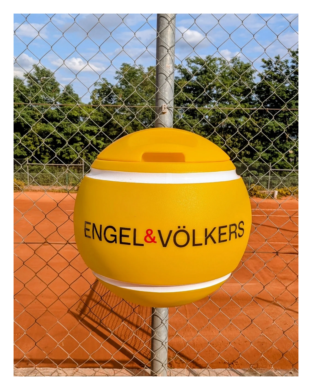 Gelber tennisballförmiger Mülleimer von Engel & Völkers auf einem Sandplatz